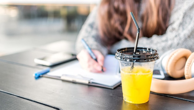 Primo piano di succo d'arancia fresco in bicchiere di plastica con coperchio e cannuccia sulla scrivania dello studente