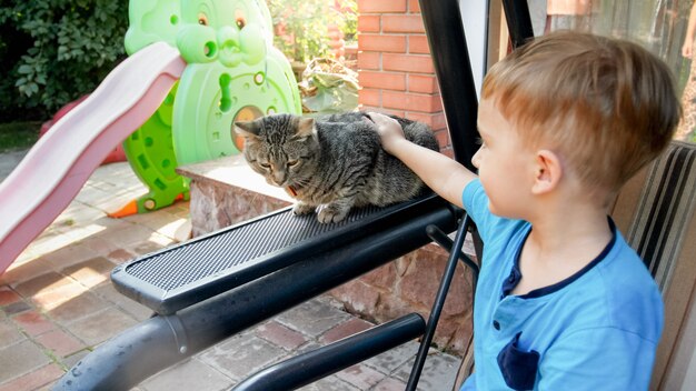 Primo piano di un adorabile bambino di 3 anni che accarezza un gatto grigio nel cortile di casa