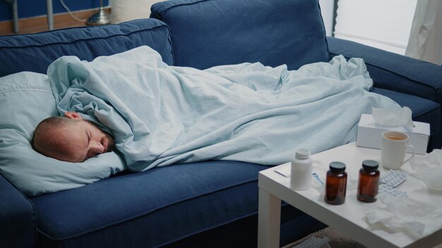 Primo piano di un adulto con sintomi influenzali sdraiato sul divano per curare il raffreddore e le malattie virali. Uomo malato che dorme e ha un trattamento di prescrizione, flaconi di pillole, capsule e termometro sul tavolo