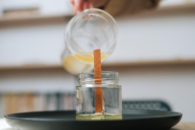 Primo piano di un'artigiana femminile irriconoscibile che versa cera fusa in un barattolo di vetro trasparente con stoppino a casa. Processo di realizzazione di candele naturali fatte a mano in officina.