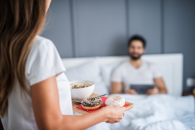 Primo piano di un'attenta giovane donna romantica porta la colazione a letto al suo ragazzo a casa o in hotel.