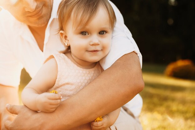 Primo piano di un bambino adorabile che guarda l'obbiettivo sorridente essendo tra le braccia di suo padre. Bambina che abbraccia con il padre all'aperto.