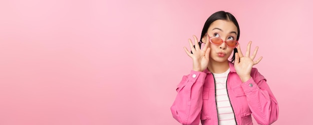 Primo piano di un bellissimo modello femminile asiatico in occhiali da sole alla moda in posa su sfondo rosa in uno spazio di copia vestito alla moda