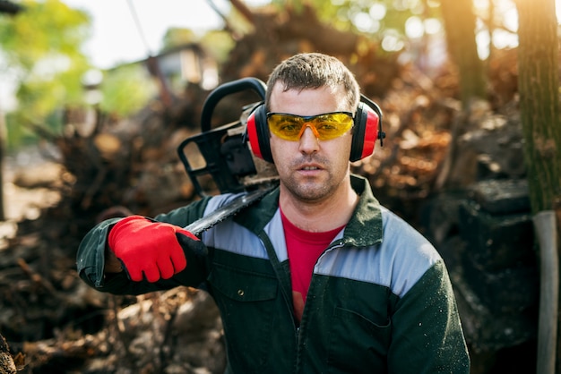 Primo piano di un boscaiolo professionista con occhiali e protezioni per le orecchie in possesso di una motosega sulla spalla pur avendo una pausa.