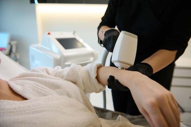 Primo piano di un estetista cosmetologo irriconoscibile che esegue una procedura di depilazione sulla mano di una donna con moderne apparecchiature laser nella clinica termale del benessere