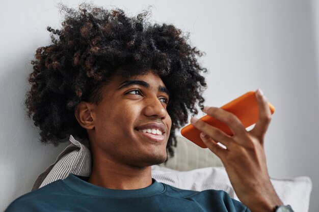 Primo piano di un giovane afro-americano che parla da smartphone sul letto e sorride, si concentra sui capelli naturali, copia spazio