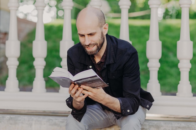 Primo piano di un giovane calvo attraente e concentrato che tiene il libro legge con gioia si gode i fine settimana trascorre il tempo libero nel giardino estivo ama leggere in un'atmosfera calma Concetto di persone e relax