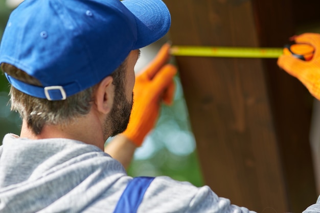 Primo piano di un giovane operaio che indossa un berretto blu e una tuta utilizzando nastro di misurazione mentre si lavora sulla costruzione di un cottage in una giornata di sole