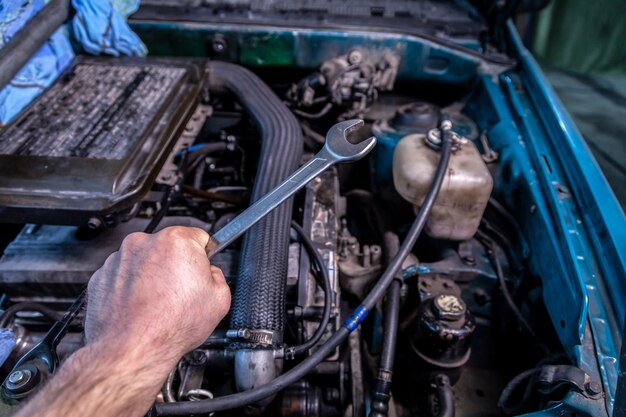 Primo piano di un giovane riparatore di auto con una chiave in mano e in procinto di riparare una macchina sotto il cofano di una macchina. concetto di riparazione auto e officina riparazioni auto