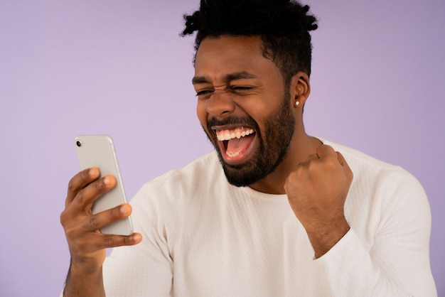 Primo piano di un giovane uomo afro che celebra la vittoria mentre guarda il suo telefono cellulare su sfondo isolato. Concetto di successo.