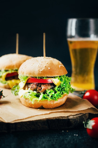 Primo piano di un hamburger con carne su un pomodoro alla griglia e verdure sullo sfondo di un bicchiere con birra su sfondo nero