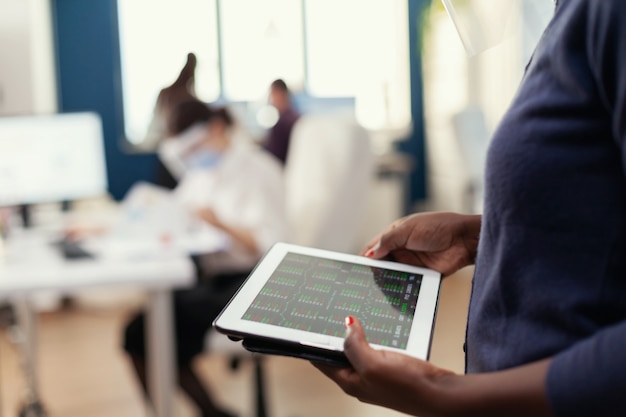 Primo piano di un imprenditore africano in un ufficio occupato utilizzando un tablet pc che analizza il grafico finanziario. Colleghi multietnici che rispettano la distanza sociale in azienda durante la pandemia globale con coronavirus.