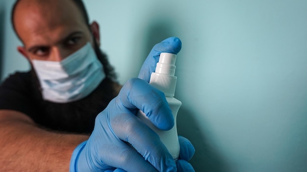 Primo piano di un maschio barbuto che indossa una maschera facciale e guanti mentre tiene in mano un flacone spray