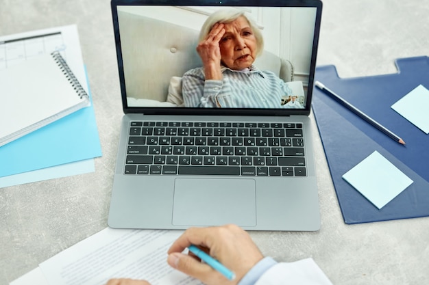 Primo piano di un medico maschio seduto al tavolo con il computer portatile e prendere appunti mentre la signora anziana descrive i sintomi