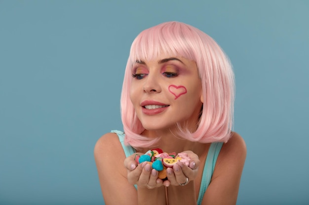 Primo piano di un modello femminile civettuolo su sfondo blu che indossa una parrucca rosa con diversi tipi di caramelle Donna piuttosto glamour capelli rosa fascino dolci stile di vita divertimento zucchero