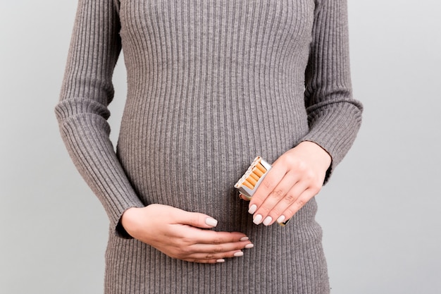 Primo piano di un pacchetto di sigarette nelle mani della donna incinta a sfondo grigio. Dipendenza da tabacco e nicotina. Il bambino è in pericolo.