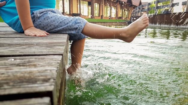 Primo piano di un ragazzino di 3 anni seduto sul molo di legno e che tiene i piedi nell'acqua del fiume. Bambino che sguazza nel lago con le gambe