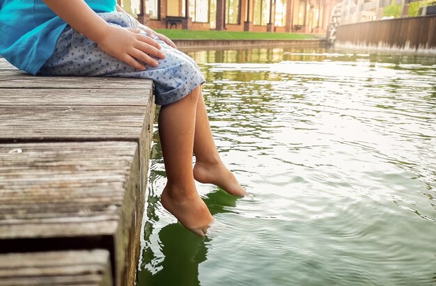 Primo piano di un ragazzino di 3 anni seduto sul molo di legno e che tiene i piedi nell'acqua del fiume. Bambino che sguazza nel lago con le gambe