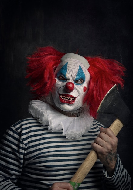 Primo piano di un spaventoso clown malvagio con capelli rossi, occhi bianchi, denti insanguinati, ascia in mano e uno sguardo minaccioso