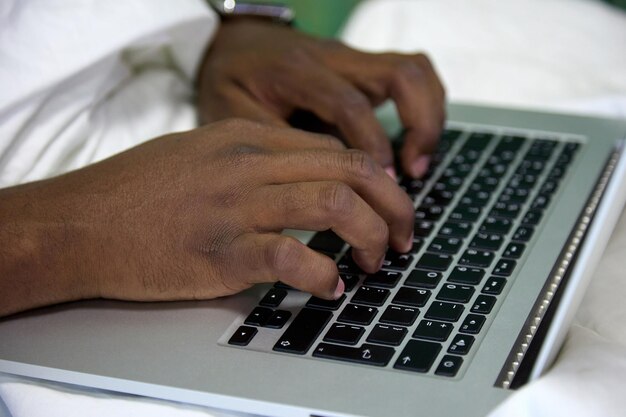 Primo piano di un uomo africano nero che scrive con le mani su un computer portatile seduto su un letto bianco