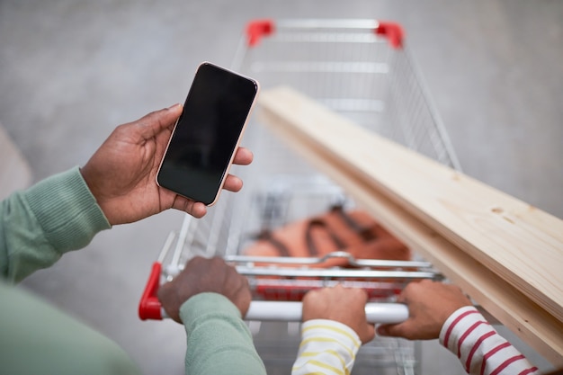 Primo piano di un uomo afroamericano irriconoscibile che tiene in mano uno smartphone con schermo vuoto mentre spinge il carrello della spesa nel supermercato, copia spazio