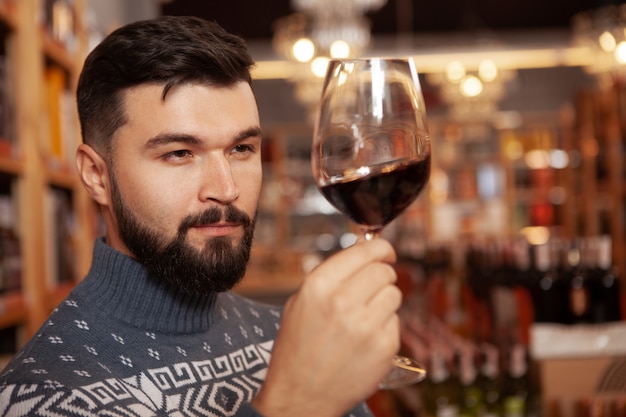 Primo piano di un uomo barbuto bello esaminando il vino rosso nel suo bicchiere, degustazione di bevande in cantina