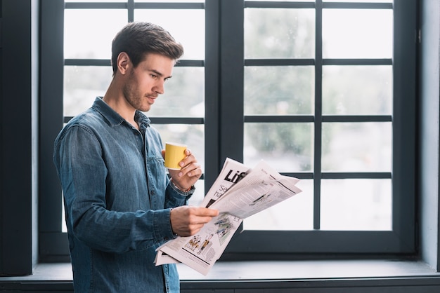 Primo piano di un uomo che tiene il giornale della lettura della tazza di caffè vicino alla finestra