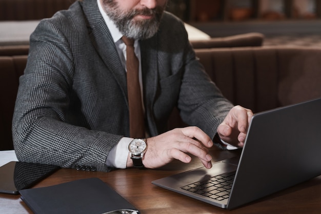 Primo piano di un uomo d'affari barbuto maturo che digita sul laptop e lavora online mentre è seduto al tavolo nella caffetteria