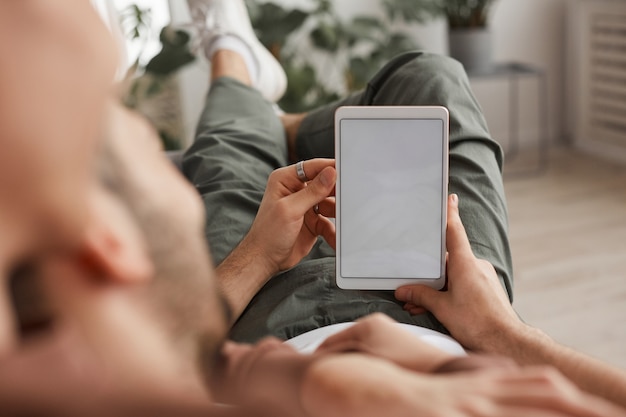 Primo piano di un uomo irriconoscibile che tiene in mano un tablet digitale con schermo bianco vuoto mentre è sdraiato sul divano a casa, copia spazio