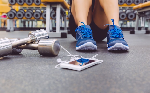 Primo piano di un uomo sportivo seduto sul pavimento del centro fitness con manubri e smartphone con auricolari in primo piano