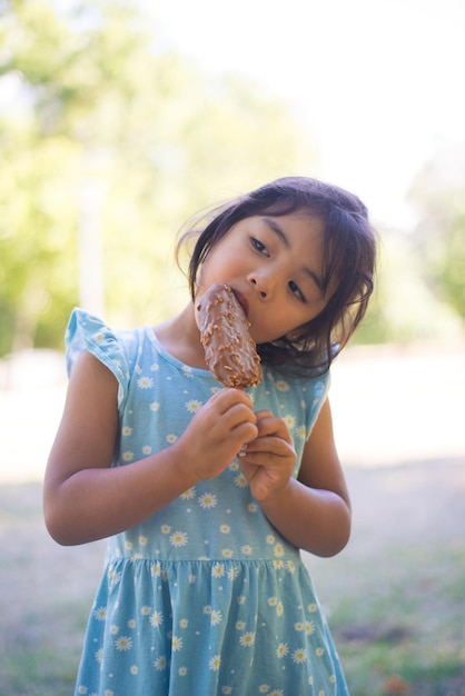Primo piano di una bambina asiatica che mangia il gelato nel parco cittadino. Bella ragazza dai capelli scuri in abito in piedi con un grande gelato sul bastone e godendosi il suo gusto. Infanzia felice, riposo estivo per il concetto di bambini