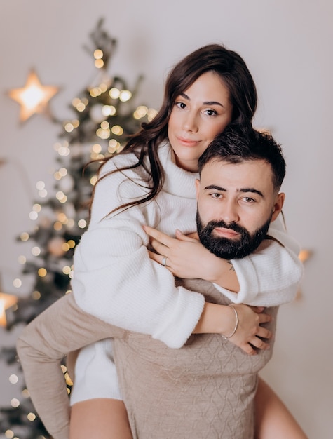 Primo piano di una bella bruna che abbraccia il suo ragazzo barbuto seduto sulla schiena sullo sfondo dell'albero di Natale