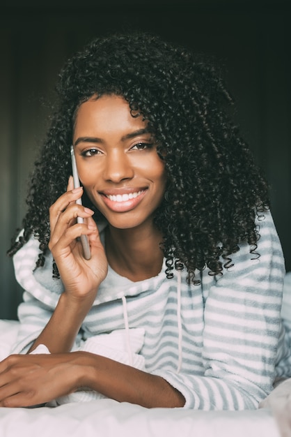 Primo piano di una bella donna nera con i capelli ricci con lo smartphone sul letto guardando la telecamera