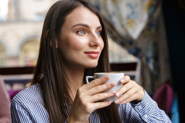 Primo piano di una bella giovane donna bruna che beve caffè mentre è seduta al bar al chiuso