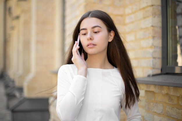 Primo piano di una bella ragazza in bianco con i capelli lunghi che parla al telefono cellulare quando si cammina per strada down