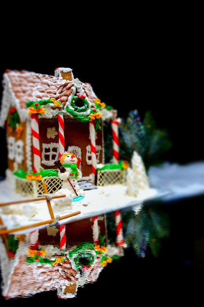 Primo piano di una casa di pan di zenzero, un rametto di albero di Natale e un pupazzo di neve di mastice di zucchero vicino al lago su sfondo scuro. Mockup per offerte stagionali e cartolina delle vacanze