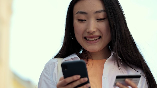 Primo piano di una donna asiatica che paga con una carta di credito per lo shopping online mentre è in piedi per strada donna che celebra un acquisto di successo