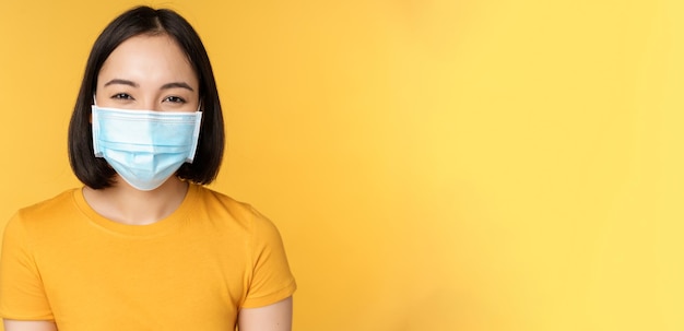 Primo piano di una donna asiatica felice sorridente che indossa una maschera medica da covid in piedi con una maglietta gialla...