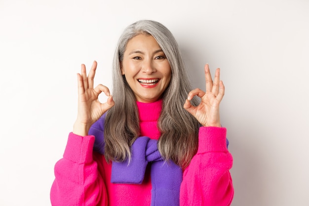 Primo piano di una donna asiatica soddisfatta con i capelli grigi, sorridente compiaciuta e che mostra segni OK, approva e apprezza il prodotto, lodando qualcosa di fantastico, sfondo bianco.