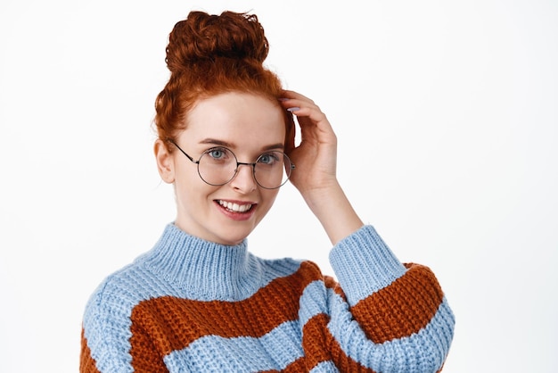 Primo piano di una donna bella e romantica con i capelli rossi pettinati in bun con gli occhiali che toccano i capelli e sorridenti che arrossiscono carino in piedi su uno sfondo bianco