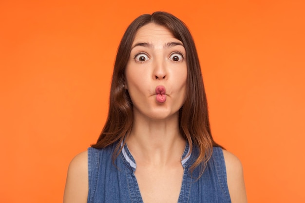 Primo piano di una donna bruna stupida divertente che fa la faccia di pesce, guardando con grandi occhi stupiti e scioccamente espressione idiota, mostrando una smorfia ridicola. tiro in studio indoor isolato su sfondo arancione