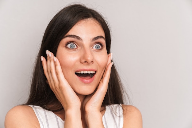 primo piano di una donna caucasica con lunghi capelli scuri che esprime sorpresa e si tocca le guance isolate su un muro grigio