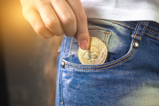 Primo piano di una donna che mette bitcoin nella tasca dei jeans, moneta di criptovaluta dorata nella foto di sfondo della tasca