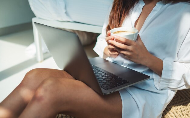 Primo piano di una donna che utilizza e che lavora al computer portatile mentre bevendo caffè e sedendosi nella camera da letto a casa