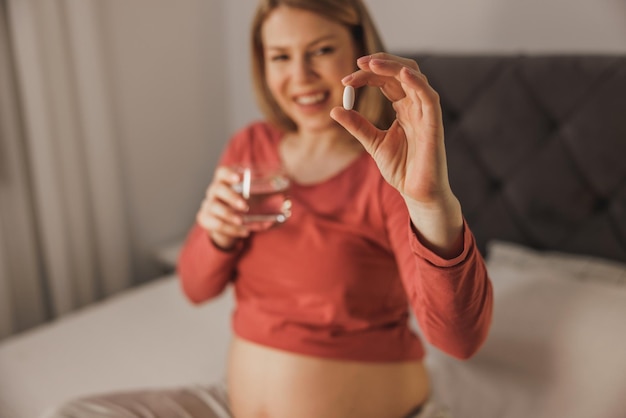 Primo piano di una donna in attesa che tiene un bicchiere con acqua e beve medicine su un letto in camera da letto.