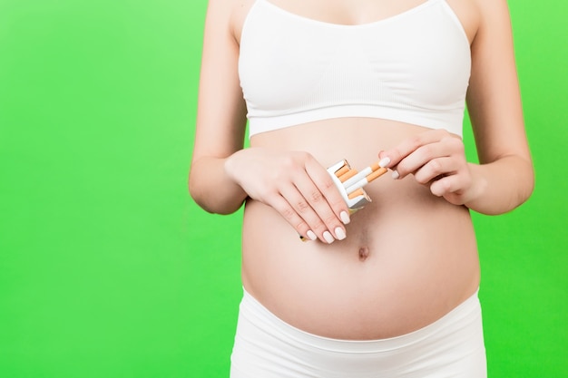Primo piano di una donna incinta in biancheria intima bianca che tiene un pacchetto di sigarette sulla superficie verde