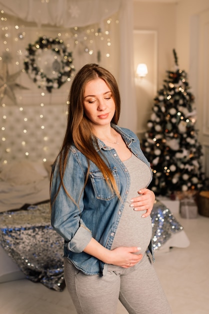 Primo piano di una donna incinta sorridente felice che si tocca la pancia sullo sfondo dell'albero di natale