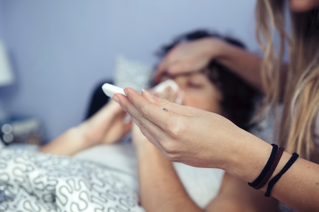 Primo piano di una donna irriconoscibile che osserva la temperatura nel termometro mentre copre la testa dell'uomo malato con il palmo della mano. La malattia e il concetto di assistenza sanitaria.