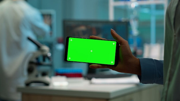 Primo piano di una donna scienziata che tiene in mano un telefono orizzontale con un modello verde in un moderno laboratorio attrezzato. Team di microbiologi che effettuano ricerche sui vaccini scrivendo su dispositivo con chiave cromatica, display isolato.