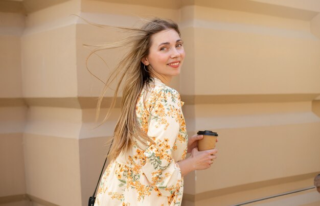 Primo piano di una donna sorridente con capelli biondi svolazzanti in un vestito che beve caffè da asporto sullo sfondo di un muro beige beige